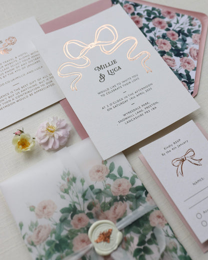 Bow Wedding Invitation featuring a large Rose Gold Bow Design, foiled onto  thick textured card with couples names and information printed in a soft brown colour. Paired with dusky pink envelope with english rose garden floral envelope liner. Laid out flat lay style with Details card and RSVP and wild roses. Designed by Fancy Folk Wedding Stationery