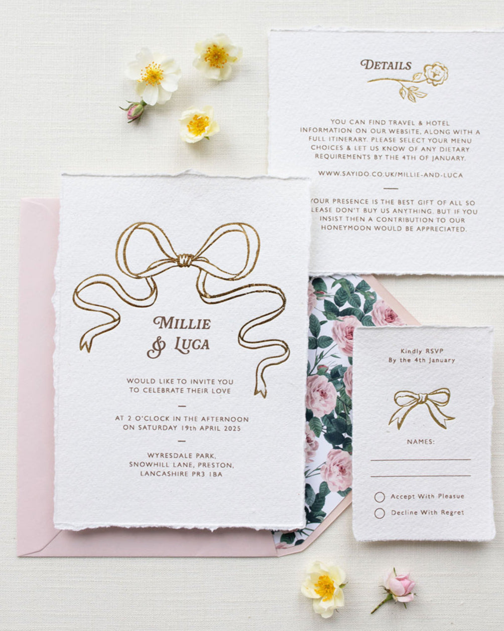 Bridgerton Inspired Gold Foil Bow design wedding invitation suite. Blush pink envelope paired with floral rose garden envelope liner. Matching Details card and RSVP cards. Printed on a luxurious and tactile handmade cotton paper. Flat lay styled  with wild roses. Designed by Fancy Folk Weddings