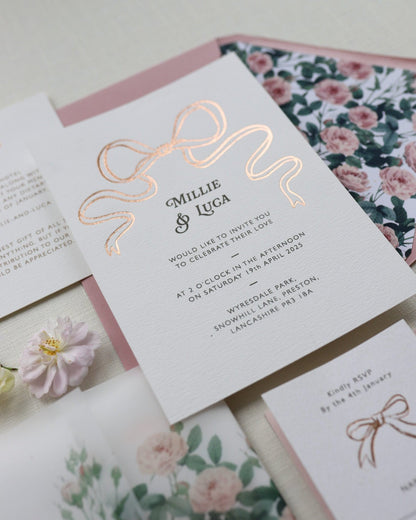 Bow Wedding Invitation featuring a large Gold Bow Design, foiled onto  thick textured card with couples names and information printed in a soft brown colour. Paired with dusky pink envelope with english rose garden floral envelope liner. Laid out flat lay style with Details card and RSVP and wild roses. Designed by Fancy Folk Wedding Stationery