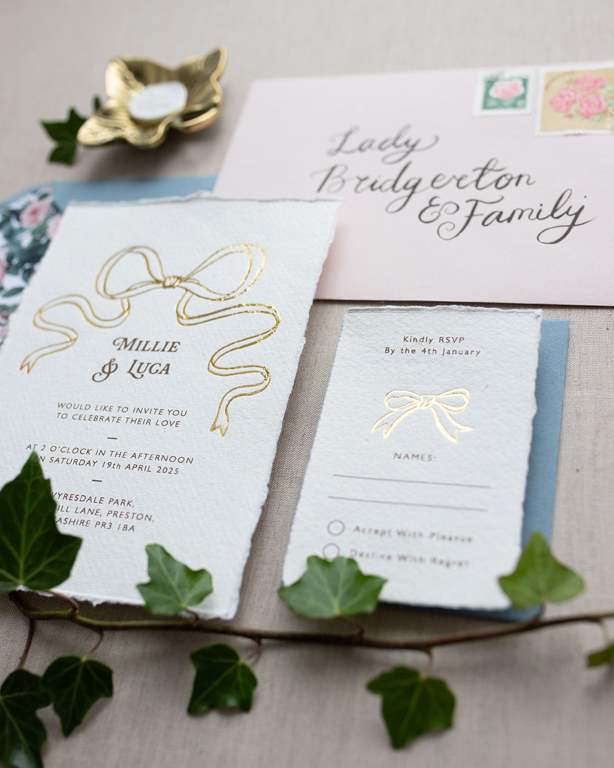 Bridgerton Inspired Gold Foil Bow design wedding invitation suite. Dusky blue coloured envelope paired with floral rose garden envelope liner. Matching RSVP card and calligraphy envelope addressing reads "Lady Bridgerton and Family". Printed on a luxurious and tactile handmade cotton paper. Flat lay styled  with Ivy. Designed by Fancy Folk Weddings