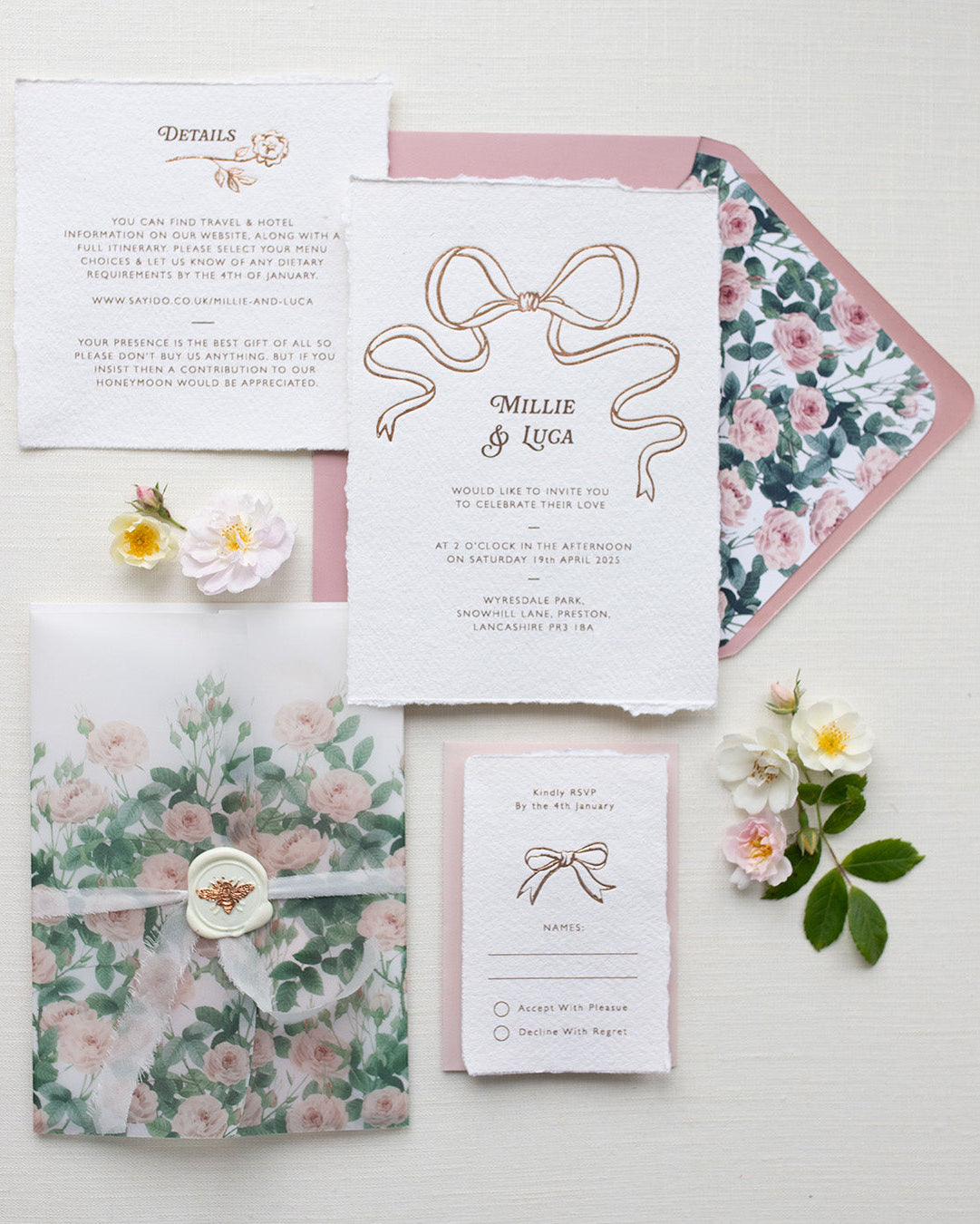 Bridgerton Inspired Rose Gold Foil Bow design wedding invitation suite. Dusky pink envelope paired with floral rose garden envelope liner. Matching Details card and RSVP cards. Printed on a luxurious and tactile handmade cotton paper. All beautifully wrapped up in a semi transparent rose print vellum wrap, silk ribbon and bee wax seal. Flat lay styled  with wild roses. Designed by Fancy Folk Weddings