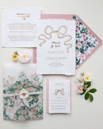 Bridgerton Inspired Rose Gold Foil Bow design wedding invitation suite. Dusky pink envelope paired with floral rose garden envelope liner. Matching Details card and RSVP cards. Printed on a luxurious and tactile handmade cotton paper. All beautifully wrapped up in a semi transparent rose print vellum wrap, silk ribbon and bee wax seal. Flat lay styled  with wild roses. Designed by Fancy Folk Weddings
