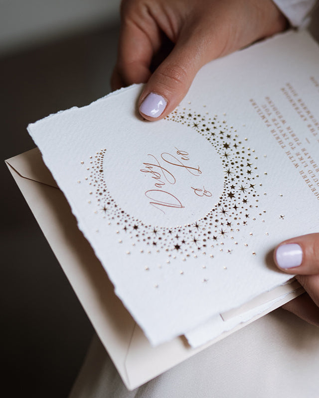 Celestial themed wedding invitation being opened by a lady. A truly magical crescent moon design made up of lots of little stars is hot foiled in gold on to tactile handmade paper with a torn romantic edge. FancyFolk Design