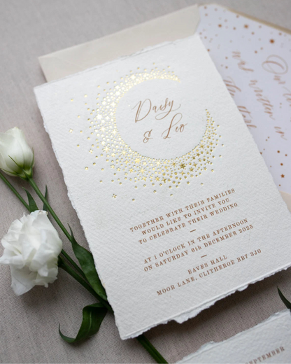 Celestial themed wedding invitation and almond coloured envelope. A truly magical crescent moon design made up of lots of little stars is hot foiled in gold on to tactile handmade paper with a torn romantic edge. The gold foil sparkles in the light. Luxury envelope liner reads "Our love was written I the stars". FancyFolk Design