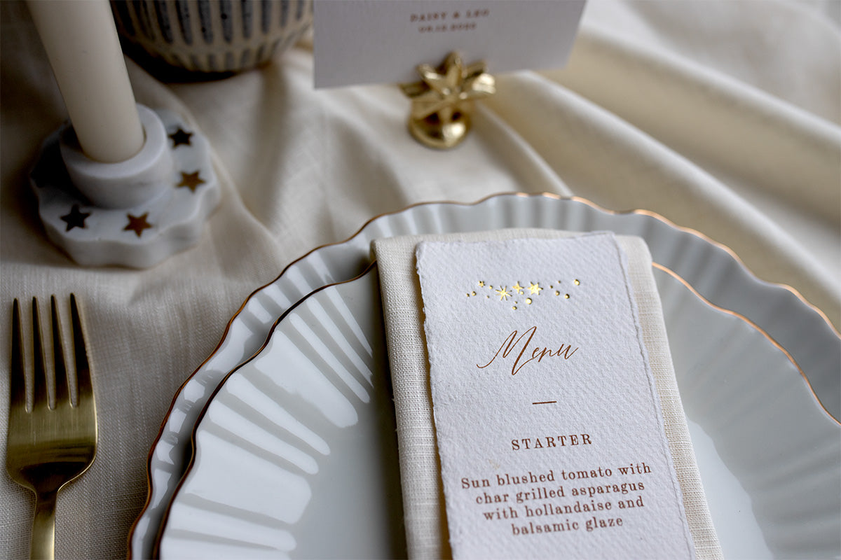 Close up of a Celestial Wedding menu. Featuring a little constellation of gold foil stars at the top and printed menu information below. Styled placed on top of a neutral napkin on beautiful crockery with gold gilding. This menu is foiled not handmade Indian cotton paper with hand torn edges for a really romantic tactile feel. Fancy Folk