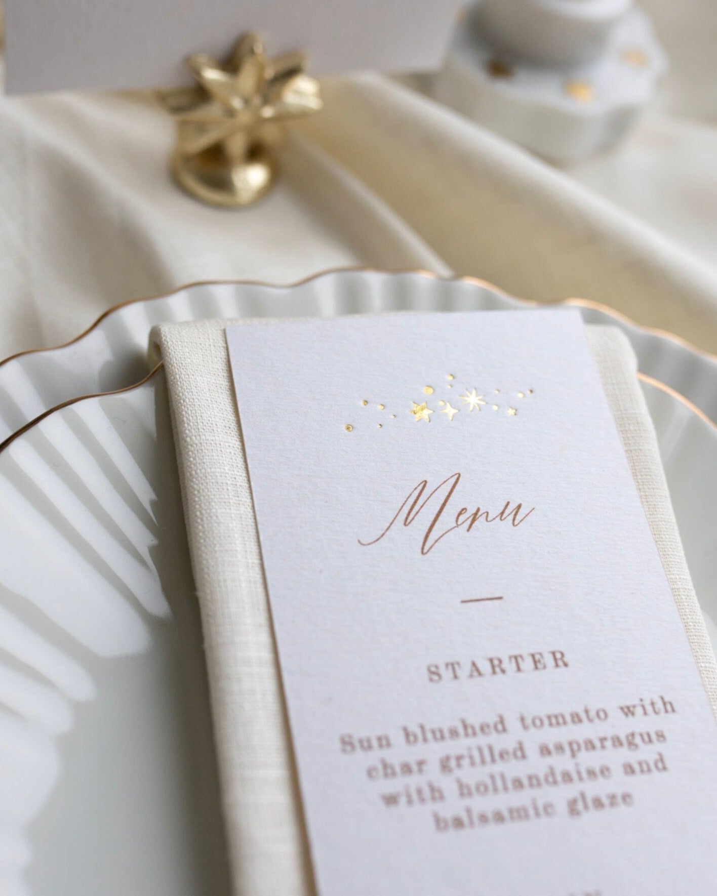 Close up of a Celestial Wedding menu, featuring a little constellation of gold foil stars at the top and printed menu information below. Styled placed on top of a neutral napkin on beautiful crockery with gold gilding. This menu is foiled not thick textured card. Fancy Folk