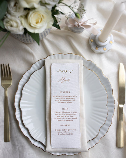 Celestial Wedding menu, featuring a little constellation of gold foil stars at the top and printed menu information below. Styled placed on top of a neutral napkin on beautiful crockery with gold gilding. This menu is foiled not handmade Indian cotton paper with hand torn edges for a really romantic tactile feel. Fancy Folk