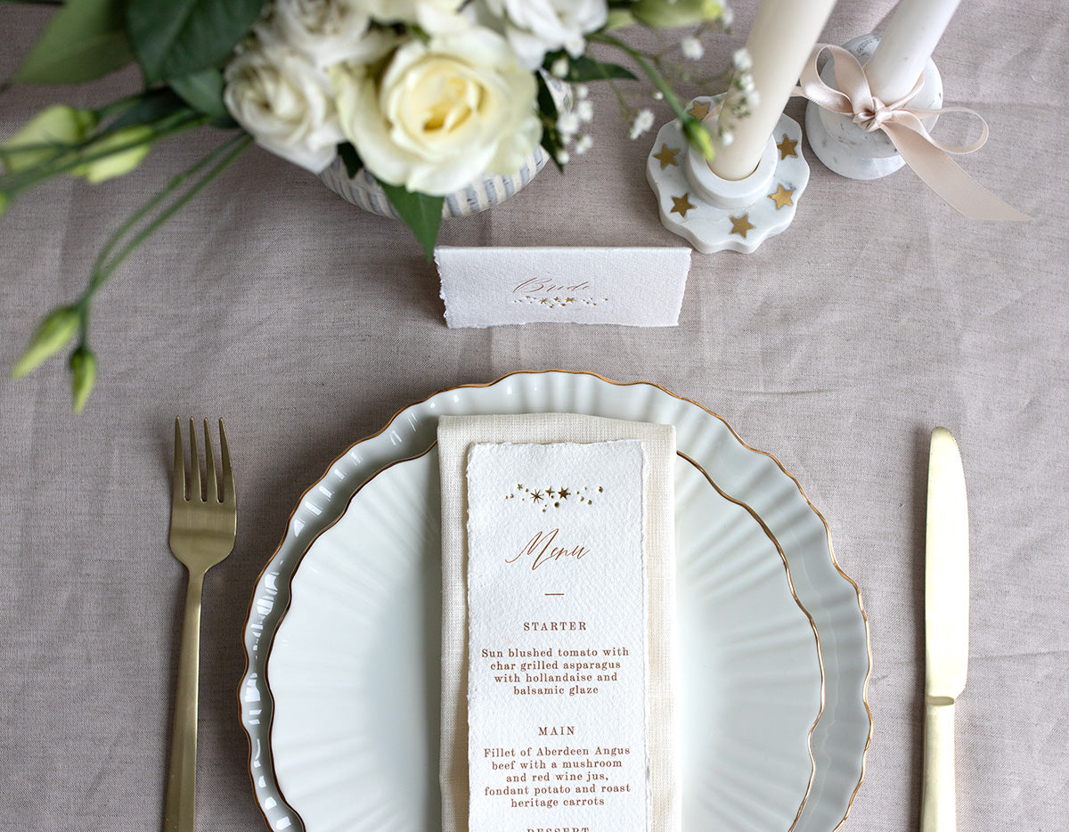Celestial Wedding menu, featuring a little constellation of gold foil stars at the top and printed menu information below. Styled placed on top of a neutral napkin on beautiful crockery with gold gilding. This menu is foiled not handmade Indian cotton paper with hand torn edges for a really romantic tactile feel. Fancy Folk