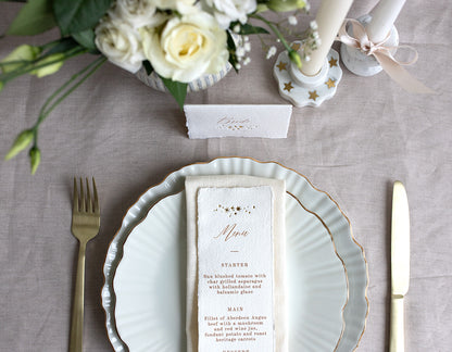 Celestial Wedding menu, featuring a little constellation of gold foil stars at the top and printed menu information below. Styled placed on top of a neutral napkin on beautiful crockery with gold gilding. This menu is foiled not handmade Indian cotton paper with hand torn edges for a really romantic tactile feel. Fancy Folk