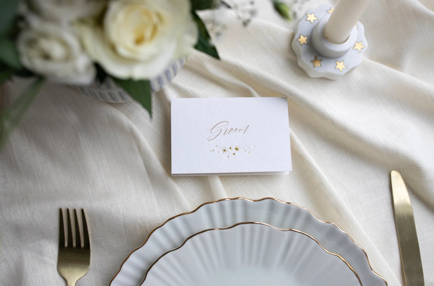Celestial wedding place card. Gold foil star constellation with printed name above, reads "Groom". Foiled onto thick textured card.. Styled with white florals and neutral linens. Fancy Folk