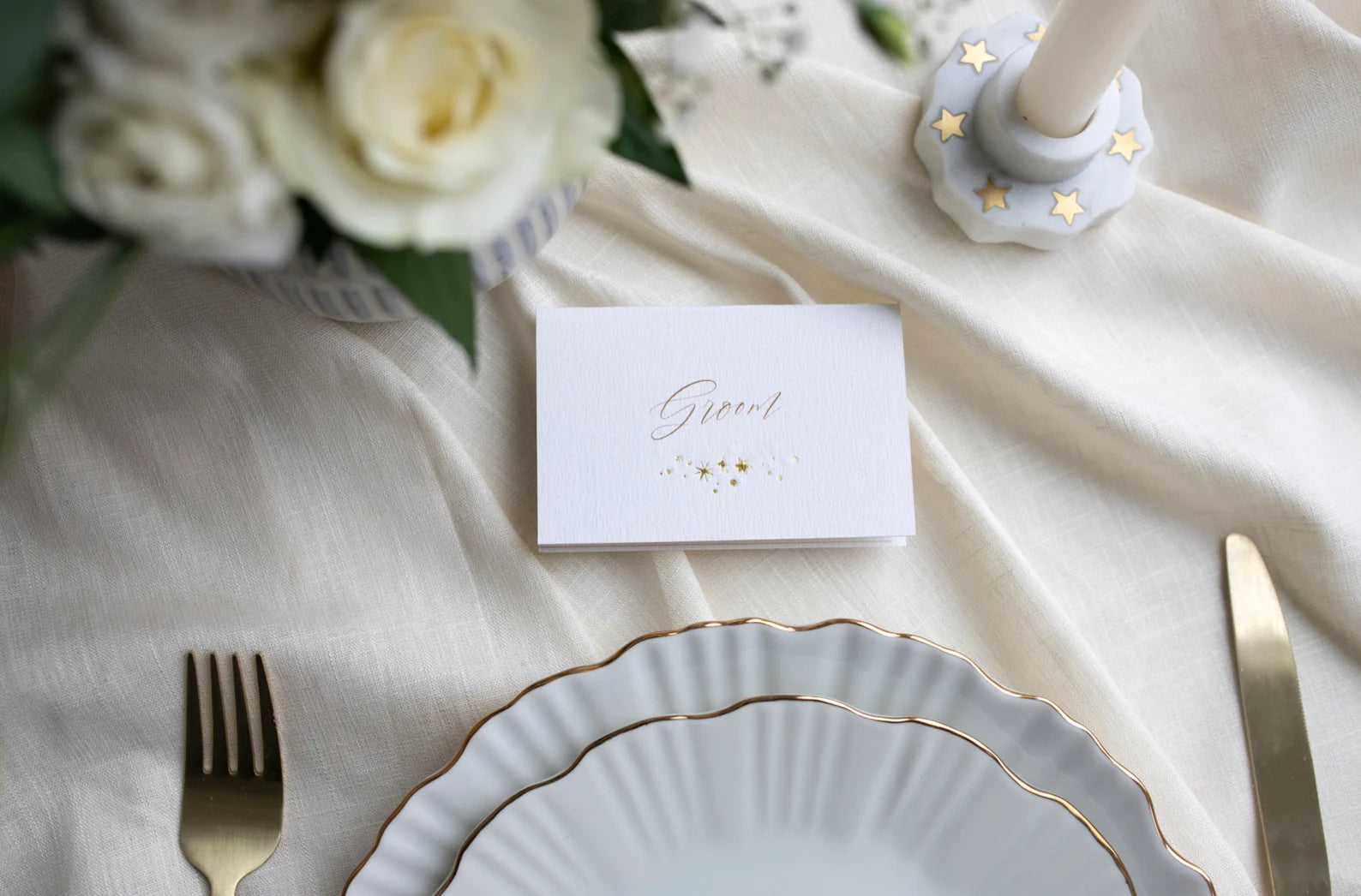 Celestial wedding place card. Gold foil star constellation with printed name above, reads "Groom". Foiled onto thick textured card.. Styled with white florals and neutral linens. Fancy Folk