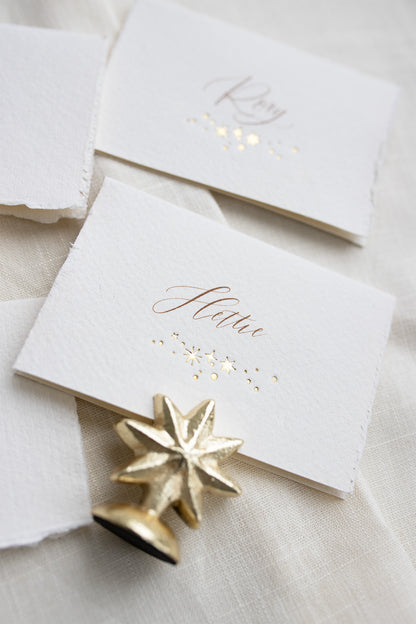 Celestial wedding place card. Gold foil star constellation with printed name above, reads "Hettie". Foiled onto handmade paper with a beautiful romantic hand torn edge. Styled with  neutral linens and gold star shaped card holder. Fancy Folk