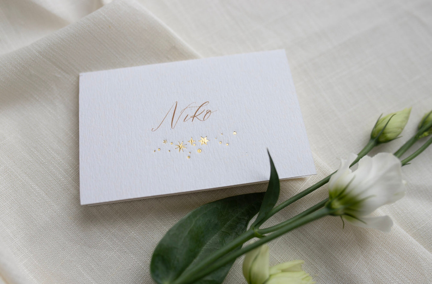 Celestial wedding place card. Gold foil star constellation with printed name above, reads "Niko". Foiled onto thick textured card.. Styled with white florals and neutral linens. Fancy Folk