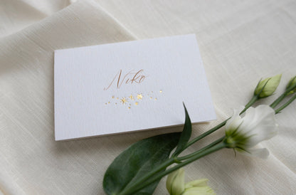 Celestial wedding place card. Gold foil star constellation with printed name above, reads "Niko". Foiled onto thick textured card.. Styled with white florals and neutral linens. Fancy Folk