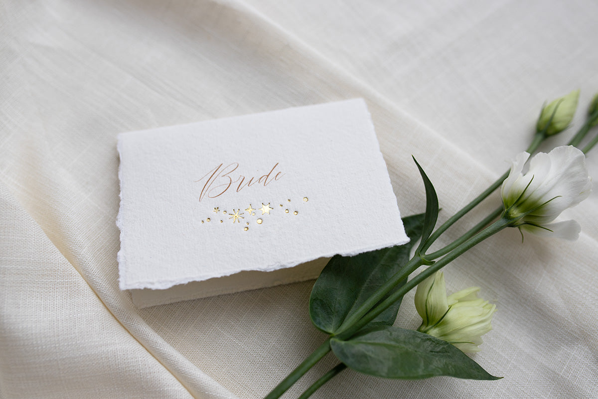 Celestial wedding place card. Gold foil star constellation with printed name above, reads "Bride". Foiled onto handmade paper with a beautiful romantic hand torn edge. Styled with white florals and neutral linens. Fancy Folk