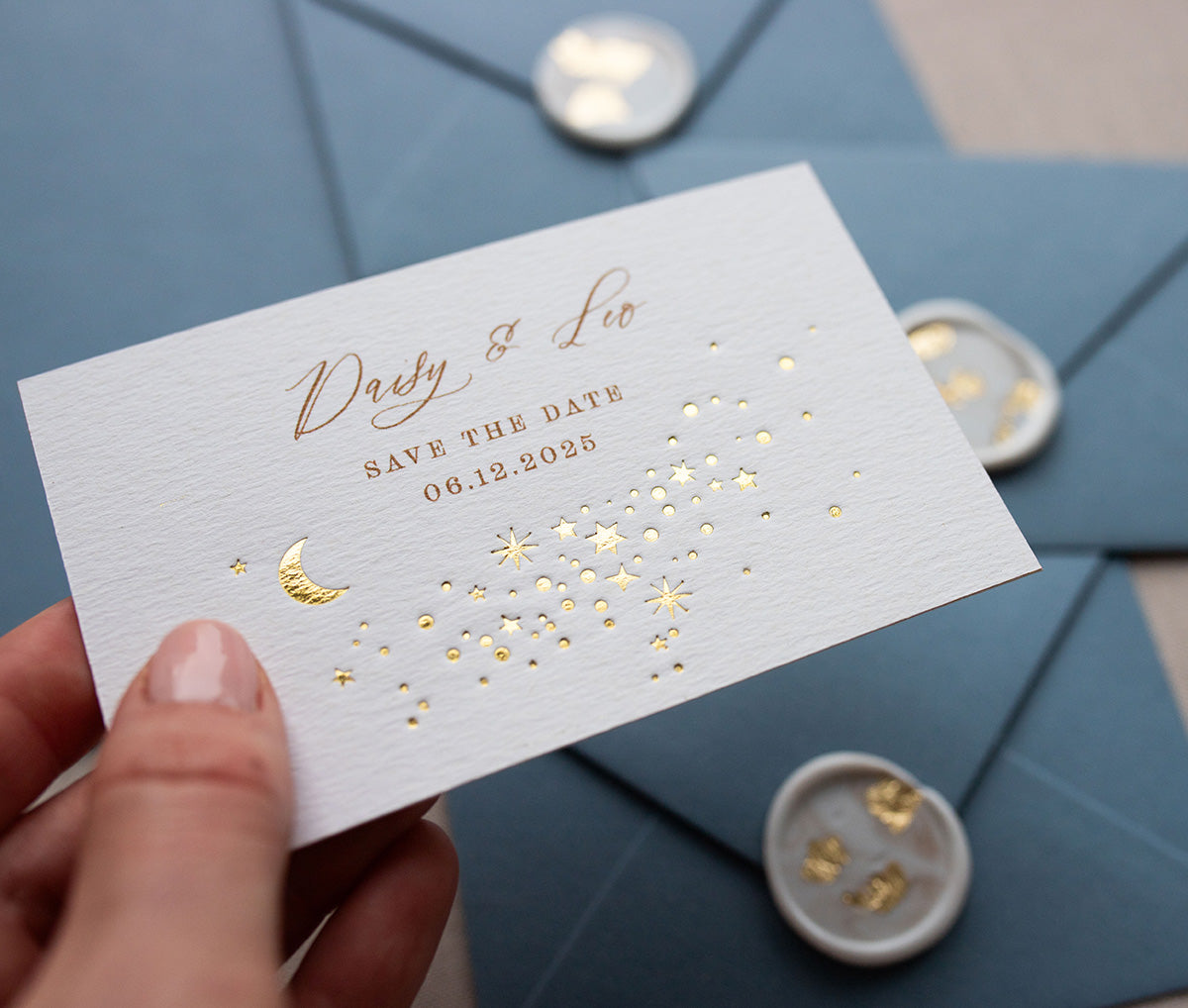 A Celestial Wedding Save the date card with gold stars and mood design. Stars are scattered across the card and  foiled in gold onto thick textured off white card. With text printed above reading "Daisy & Leo Save the Date " styled with dusty blue coloured envelopes and white wax seals with flakes of gold leaf embedded in them for a luxury finish. Fancy Folk Design.