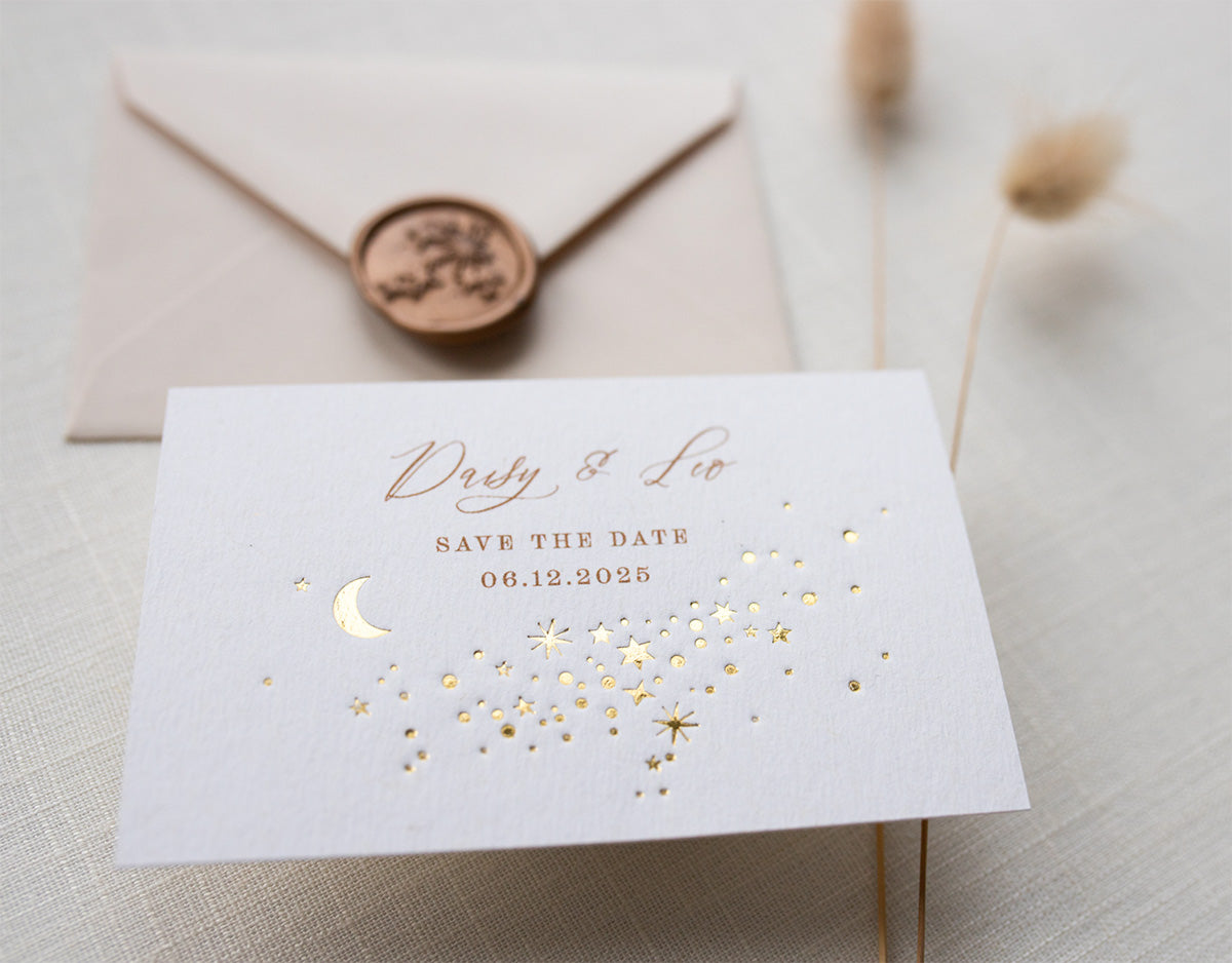 A Celestial Wedding Save the date card with gold stars and mood design. Stars are scattered across the card and  foiled in gold onto thick textured off white card. With text printed above reading "Daisy & Leo Save the Date " styled with neutral almond coloured envelope. Fancy Folk Design.