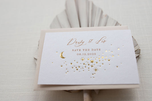 A Celestial Wedding Save the date card with gold stars and mood design. Stars are scattered across the card and  foiled in gold onto thick textured off white card. With text printed above reading "Daisy & Leo Save the Date " styled with neutral almond coloured envelope. Fancy Folk Design.