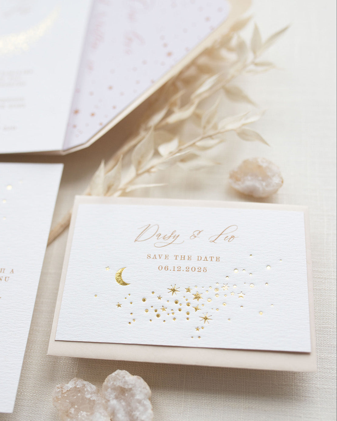 A Celestial Wedding Save the date card with gold stars and mood design. Stars are scattered across the card and  foiled in gold onto thick textured off white card. With text printed above reading "Daisy & Leo Save the Date " styled with neutral almond coloured envelope. Fancy Folk Design.