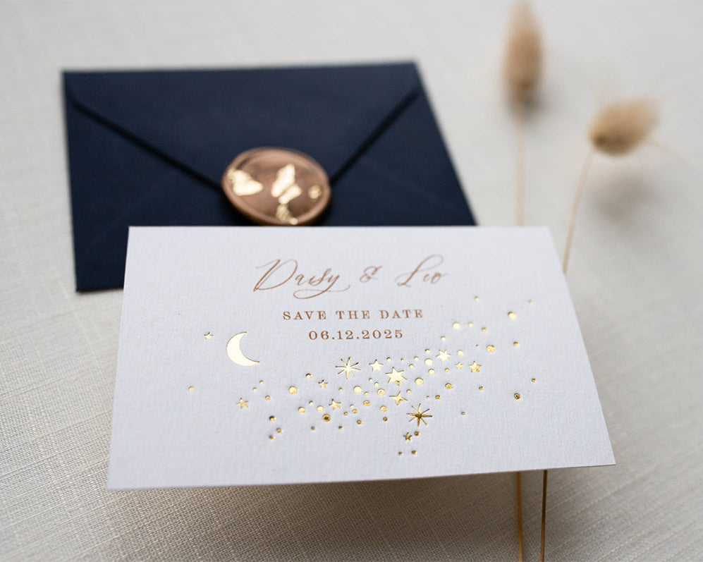 A Celestial Wedding Save the date card with gold stars and mood design. Stars are scattered across the card and  foiled in gold onto thick textured off white card. With text printed above reading "Daisy & Leo Save the Date " styled with navy blue coloured envelope. Fancy Folk Design.