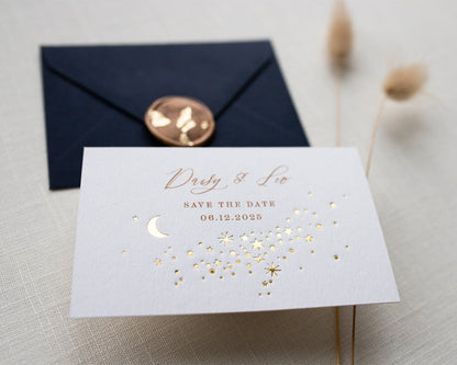 A Celestial Wedding Save the date card with gold stars and mood design. Stars are scattered across the card and  foiled in gold onto thick textured off white card. With text printed above reading "Daisy & Leo Save the Date " styled with navy blue coloured envelope. Fancy Folk Design.