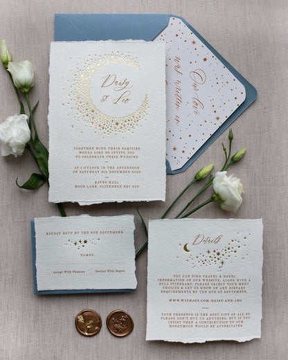 Celestial themed wedding stationery suite laid out in a flat lay format. A truly magical crescent moon design made up of lots of little stars is hot foiled in gold on to the main invitation. Smaller cluster of gold stars feature on the details card and RSVP. The invitations are foiled and printed onto tactile handmade paper with a torn romantic edge. Styled with gold wax seals, dusty blue envelopes with a luxury envelope liner which reads "Our love was written in the stars". FancyFolk Design