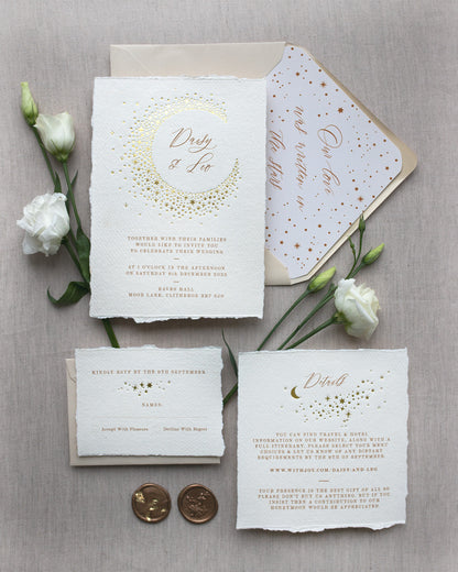 Celestial themed wedding stationery suite laid out in a flat lay format. A truly magical crescent moon design made up of lots of little stars is hot foiled in gold on to the main invitation. Smaller cluster of gold stars feature on the details card and RSVP. The invitations are foiled and printed onto tactile handmade paper with a torn romantic edge. Styled with gold wax seals, neutral envelopes with a luxury envelope liner which reads "Our love was written in the stars". FancyFolk Design