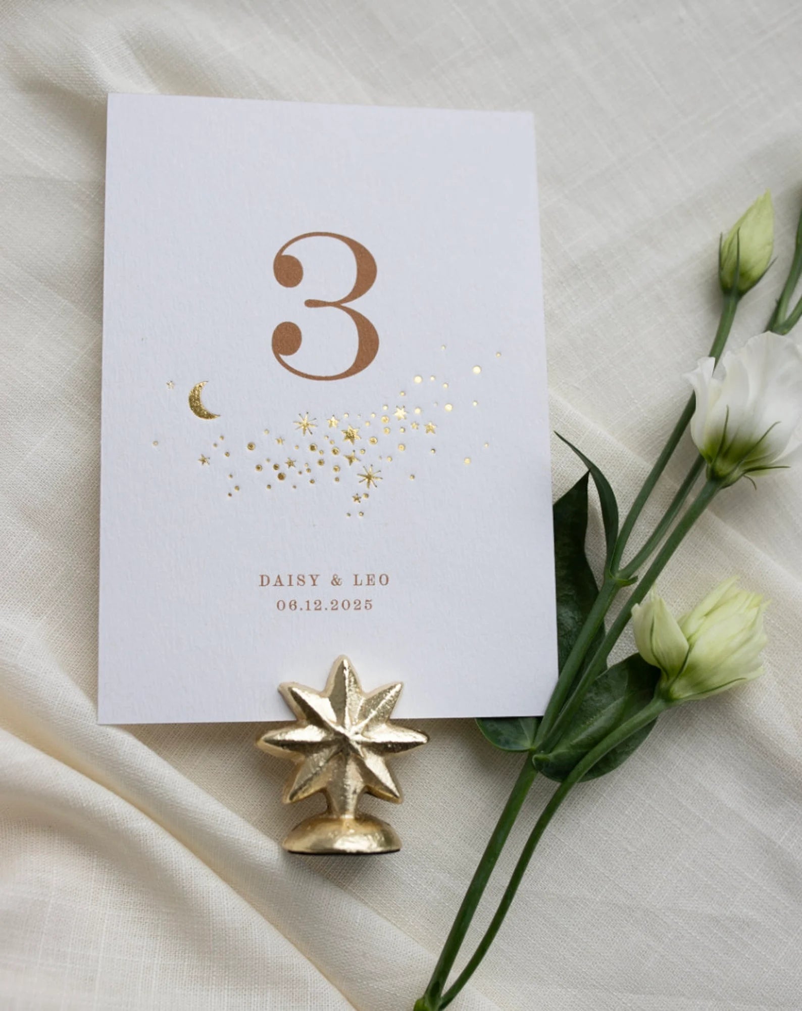 Celestial themed design featuring a scatter of gold stars and a little moon. Gold hot foiled onto thick textured card with printed number and coupled names and wedding date below. Styled with white florals, neutral linens and gold star shaped card. Fancy Folk Design