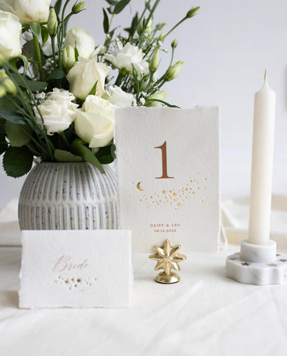 Celestial themed design featuring a scatter of gold stars and a little moon. Gold hot foiled onto handmade cotton paper with torn edges.Printed number and coupled names and wedding date below. Styled with white florals, neutral linens and gold star shaped card. Fancy Folk Design