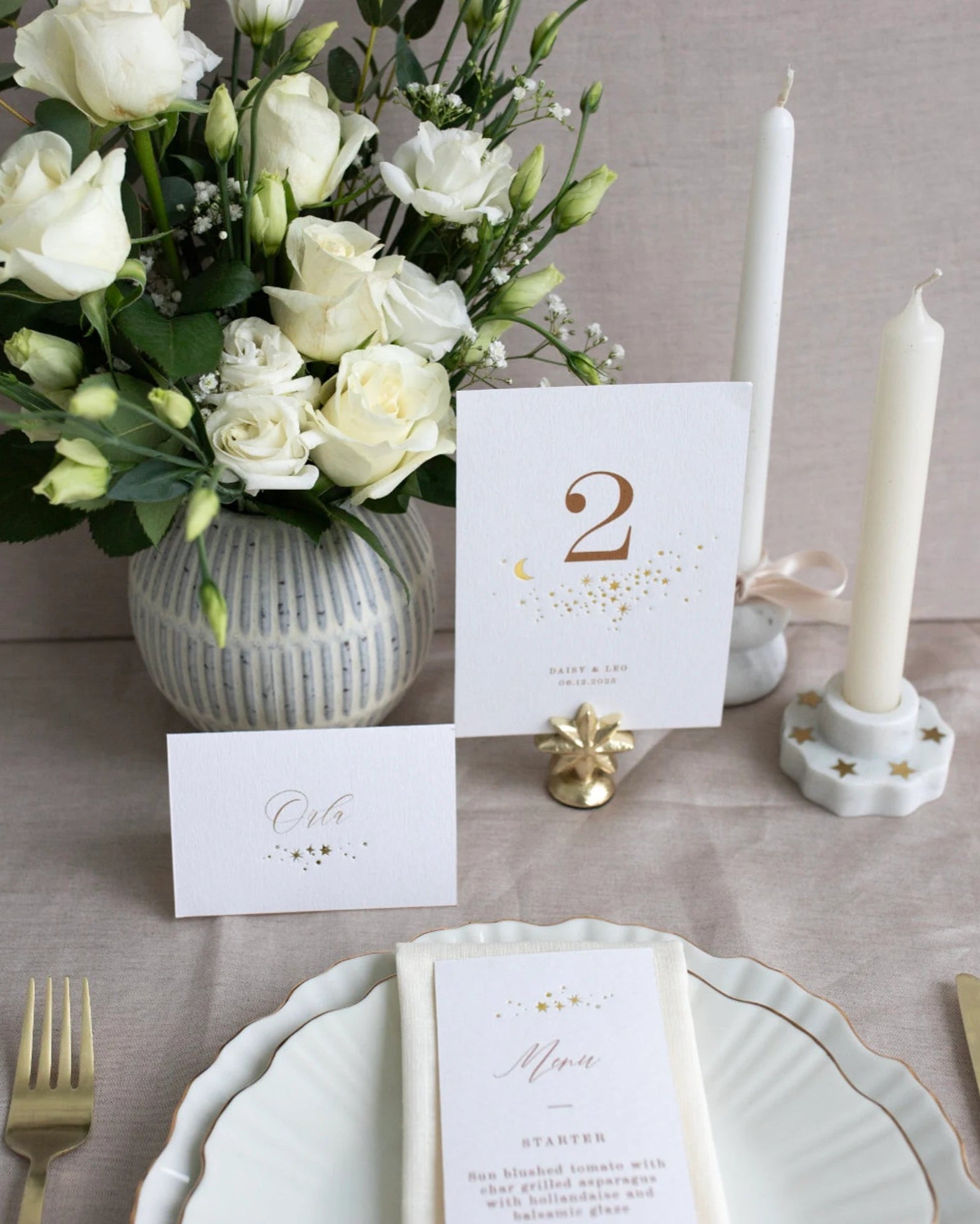 Celestial themed design featuring a scatter of gold stars and a little moon. Gold hot foiled onto thick textured card with printed number and coupled names and wedding date below. Styled with white florals, neutral linens and gold star shaped card. Photographed on a table set up with beautiful crockery and gold cutlery along with complimentary place cards and menus in the same theme.  Fancy Folk Design