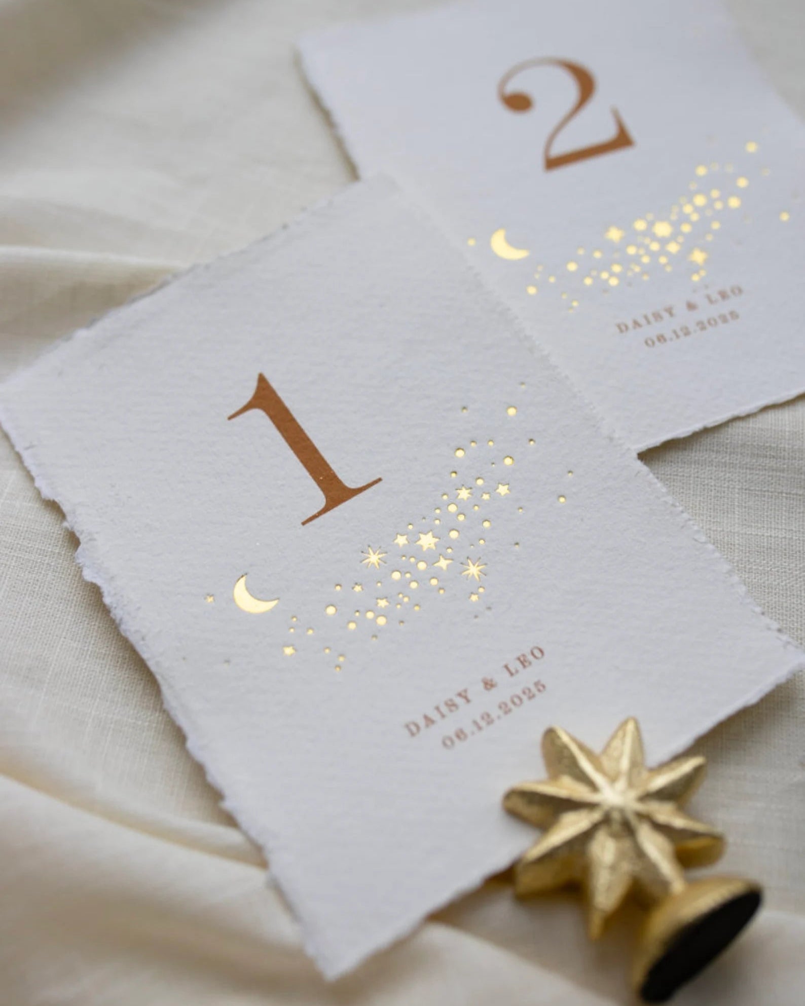 Celestial themed design featuring a scatter of gold stars and a little moon. Gold hot foiled onto handmade cotton paper with torn edges.Printed number and coupled names and wedding date below. Styled with  neutral linens and gold star shaped card. Fancy Folk Design