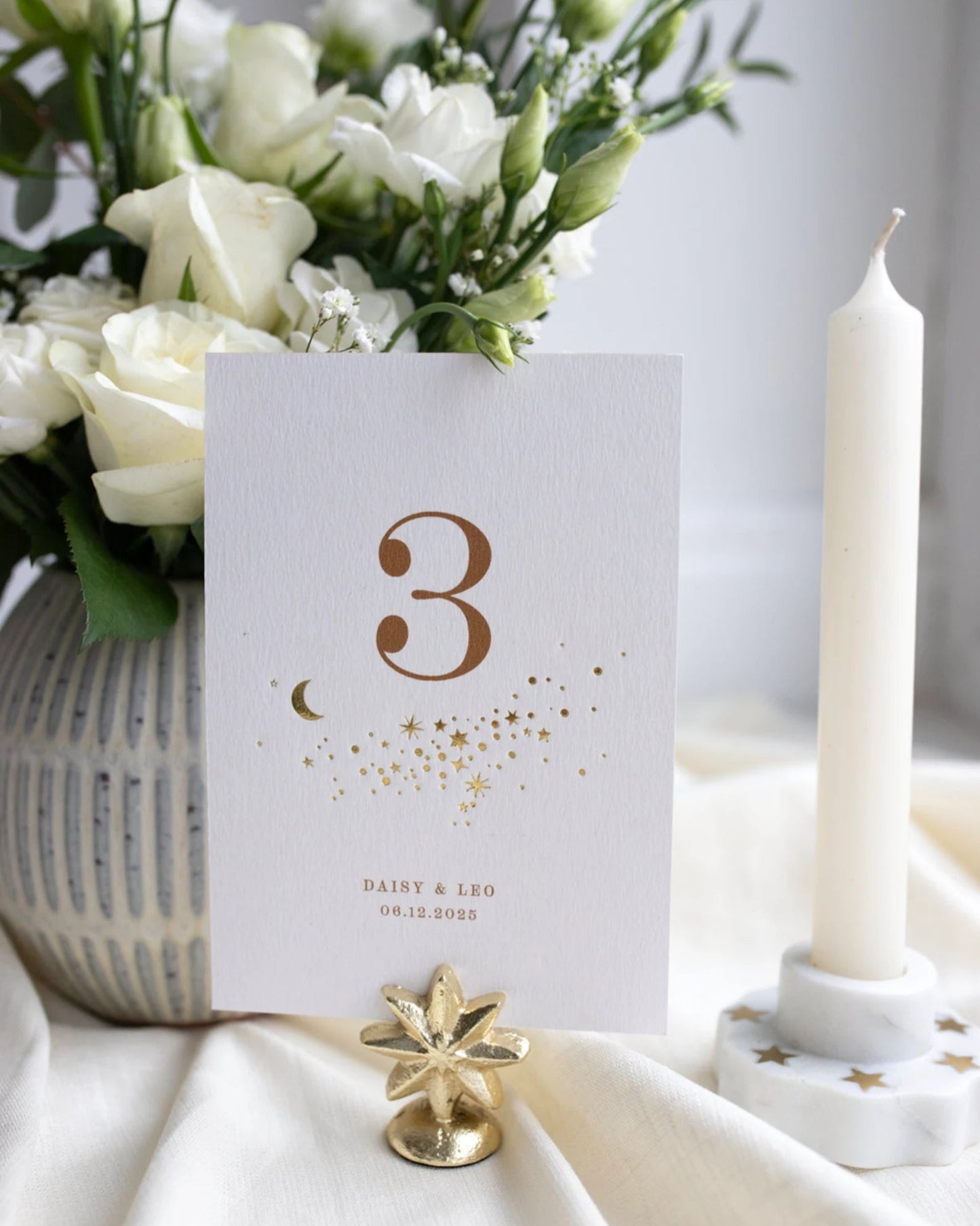 Celestial themed design featuring a scatter of gold stars and a little moon. Gold hot foiled onto thick textured card with printed number and coupled names and wedding date below. Styled with white florals, neutral linens and gold star shaped card. Fancy Folk Design