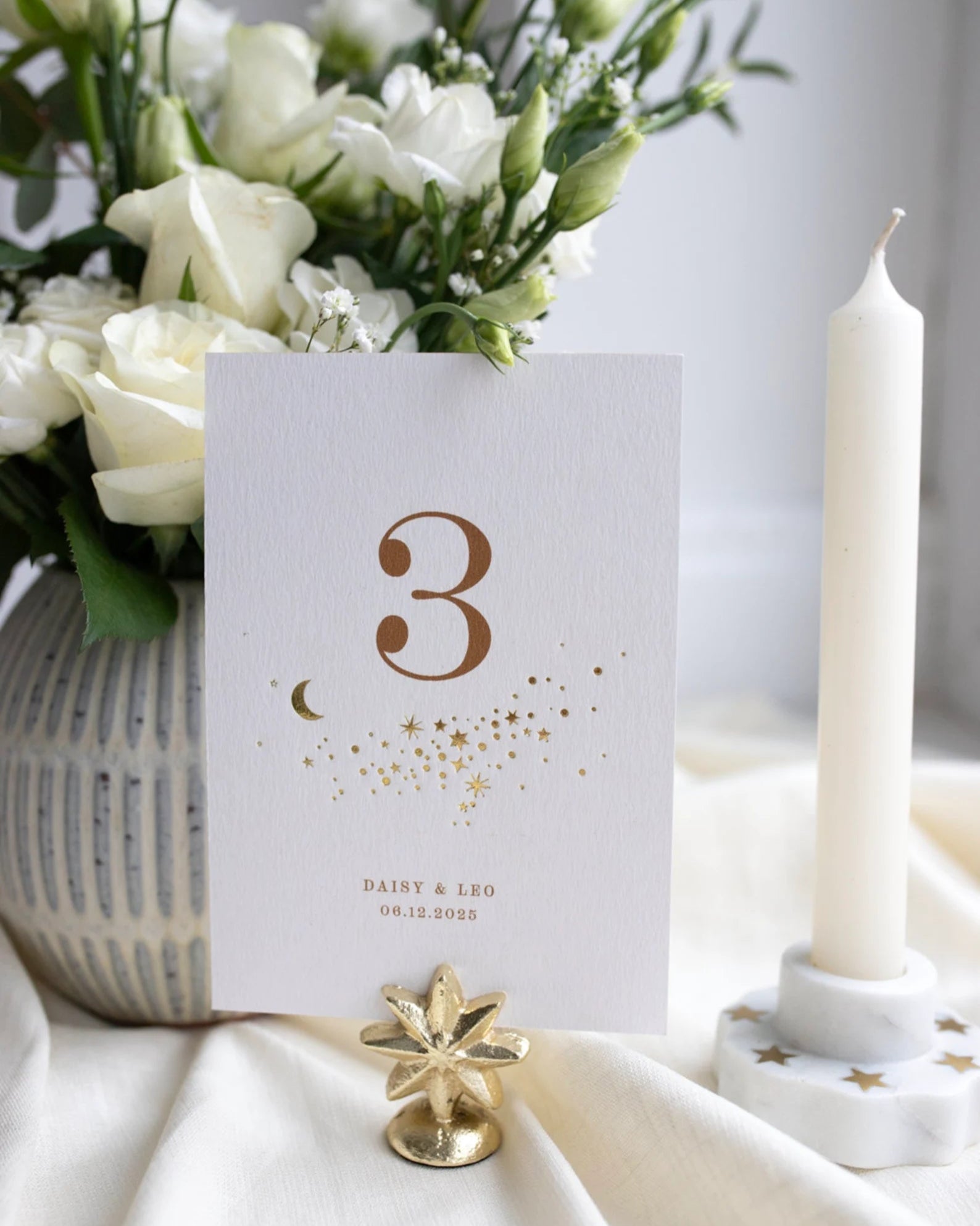 Celestial themed design featuring a scatter of gold stars and a little moon. Gold hot foiled onto thick textured card with printed number and coupled names and wedding date below. Styled with white florals, neutral linens and gold star shaped card. Fancy Folk Design