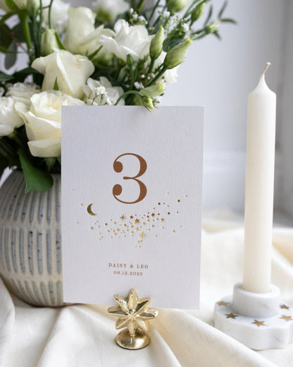 Celestial themed design featuring a scatter of gold stars and a little moon. Gold hot foiled onto thick textured card with printed number and coupled names and wedding date below. Styled with white florals, neutral linens and gold star shaped card. Fancy Folk Design