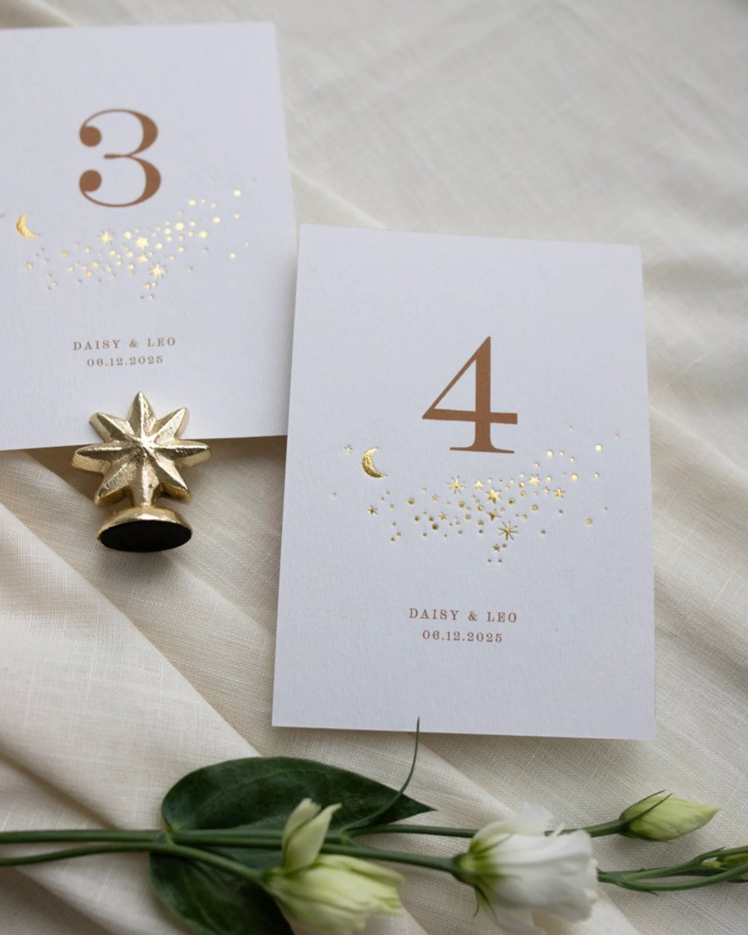 Celestial themed design featuring a scatter of gold stars and a little moon. Gold hot foiled onto thick textured card with printed number and coupled names and wedding date below. Styled with white florals, neutral linens and gold star shaped card. Fancy Folk Design