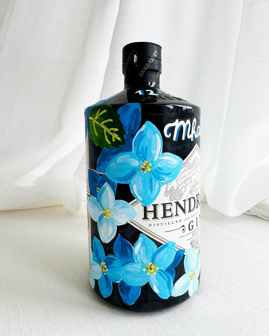 Hand painted Occasion Bottle. Blue Hydrangeas in different shades are painted all around the bottle. Calligraphy name of the recipient at the front of the bottle in white. Gold leaf accents to finish. Fancy Folk Design