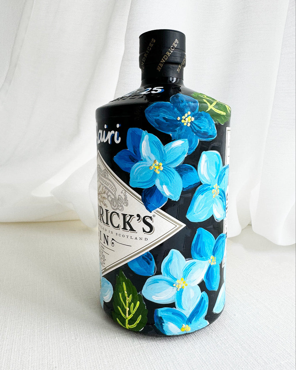 Hand painted Occasion Bottle. Blue Hydrangeas in different shades are painted all around the bottle. Calligraphy name of the recipient at the front of the bottle in white. Gold leaf accents to finish. Fancy Folk Design