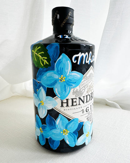 Hand painted Occasion Bottle. Blue Hydrangeas in different shades are painted all around the bottle. Calligraphy name of the recipient at the front of the bottle in white. Gold leaf accents to finish. Fancy Folk Design