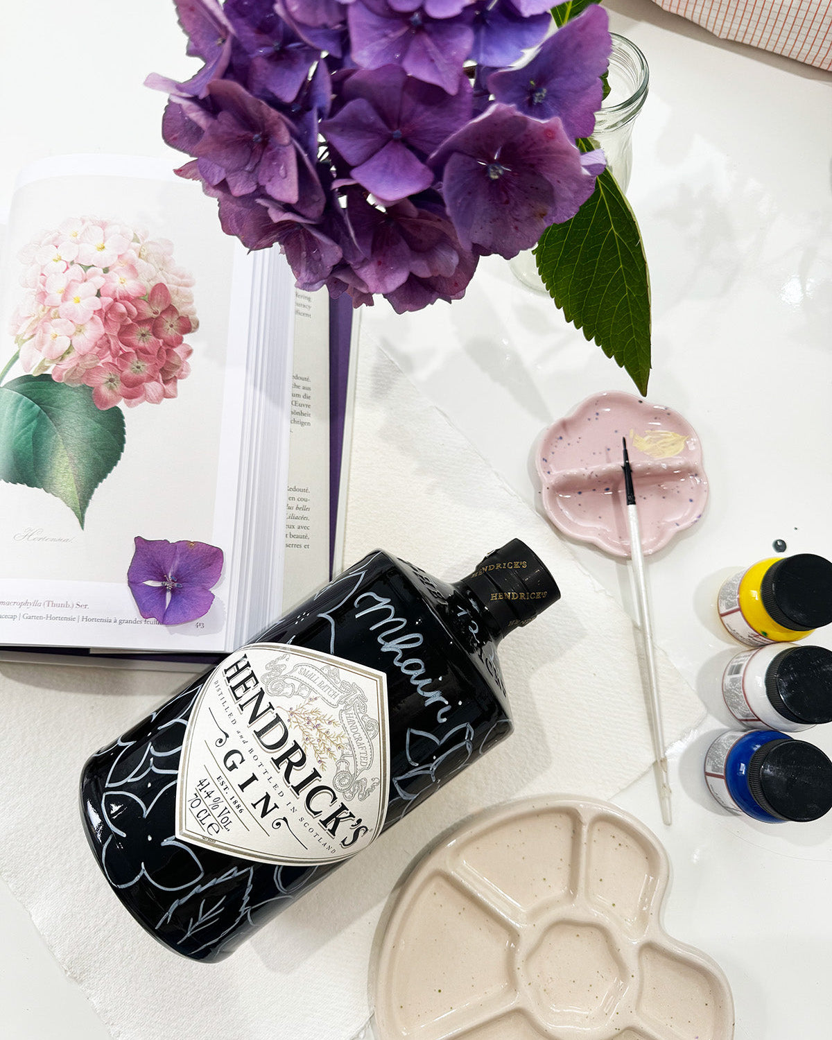 Art supplies and inspiration layer out ready to paint a Gin bottle gift. Hand painted Occasion Bottle.  Hydrangeas petals are sketched out all around the bottle ready to paint. Calligraphy name of the recipient at the front of the bottle in white. Fancy Folk Design