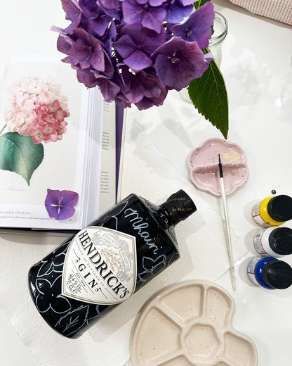 Art supplies and inspiration layer out ready to paint a Gin bottle gift. Hand painted Occasion Bottle.  Hydrangeas petals are sketched out all around the bottle ready to paint. Calligraphy name of the recipient at the front of the bottle in white. Fancy Folk Design