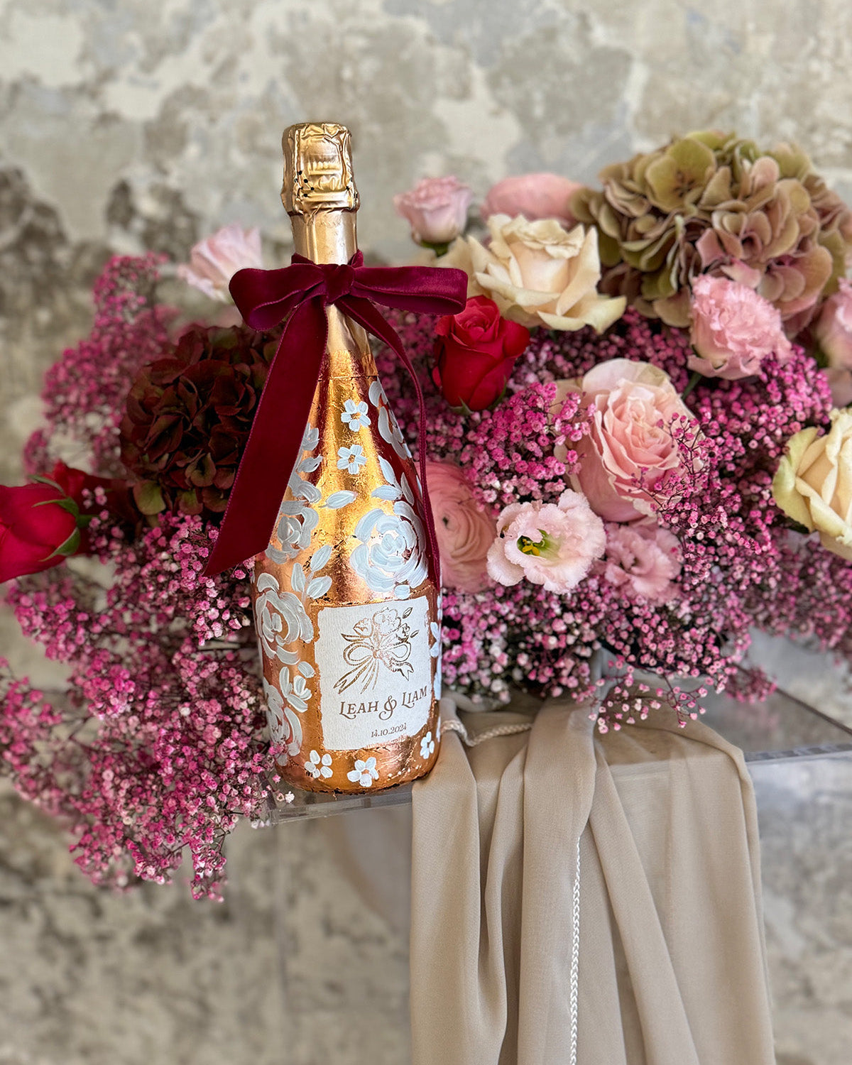 Hand painted wedding Champagne Bottle. Lux Copper leaf base with white painted roses all around the bottle. Personalise label with couples names and wedding date. Perfect for a Champagne Tower moment. Pink florals in the background. By Fancy Folk Design