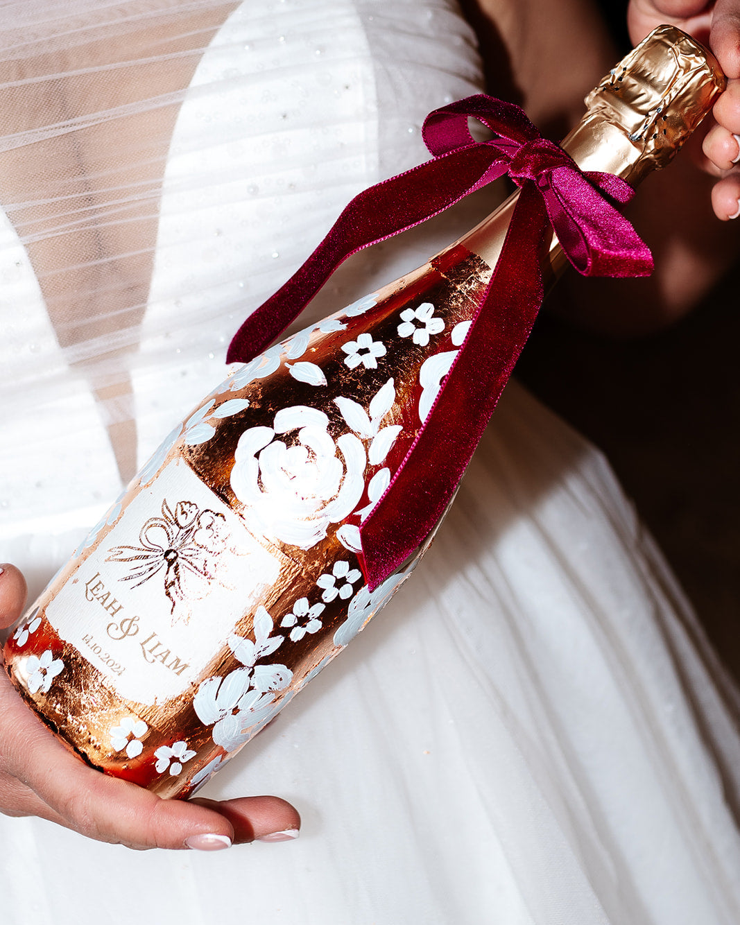 Hand painted wedding Champagne Bottle. Lux Copper leaf base with white painted roses all around the bottle. Personalise label with couples names and wedding date. Held in the brides hands. By Fancy Folk Design