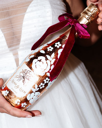 Hand painted wedding Champagne Bottle. Lux Copper leaf base with white painted roses all around the bottle. Personalise label with couples names and wedding date. Held in the brides hands. By Fancy Folk Design