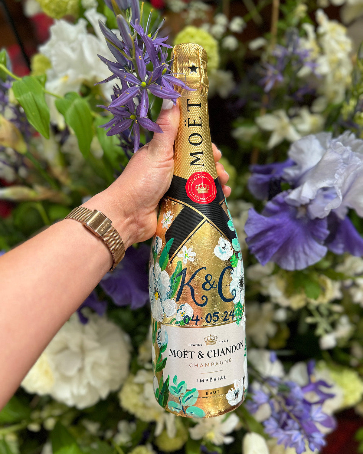 Hand painted Bottle of Moet Champagne. Perfect wedding gift with couples monogram initials and wedding date. Gold leaf base painted florals to match the bridal bouquet. Woman's hands holding the bottle. Fancy Folk Design