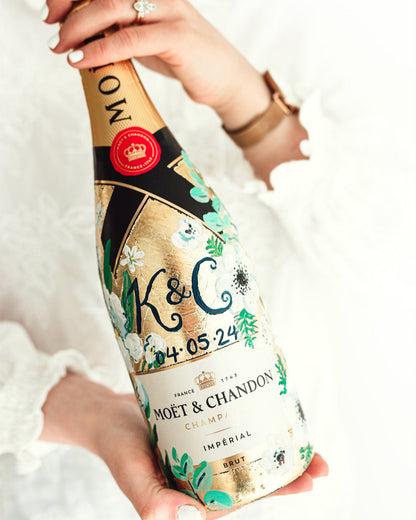 Hand painted Bottle of Moet Champagne. Perfect wedding gift with couples monogram initials and wedding date. Gold leaf base painted florals to match the bridal bouquet. Woman's hands holding the bottle. Fancy Folk Design