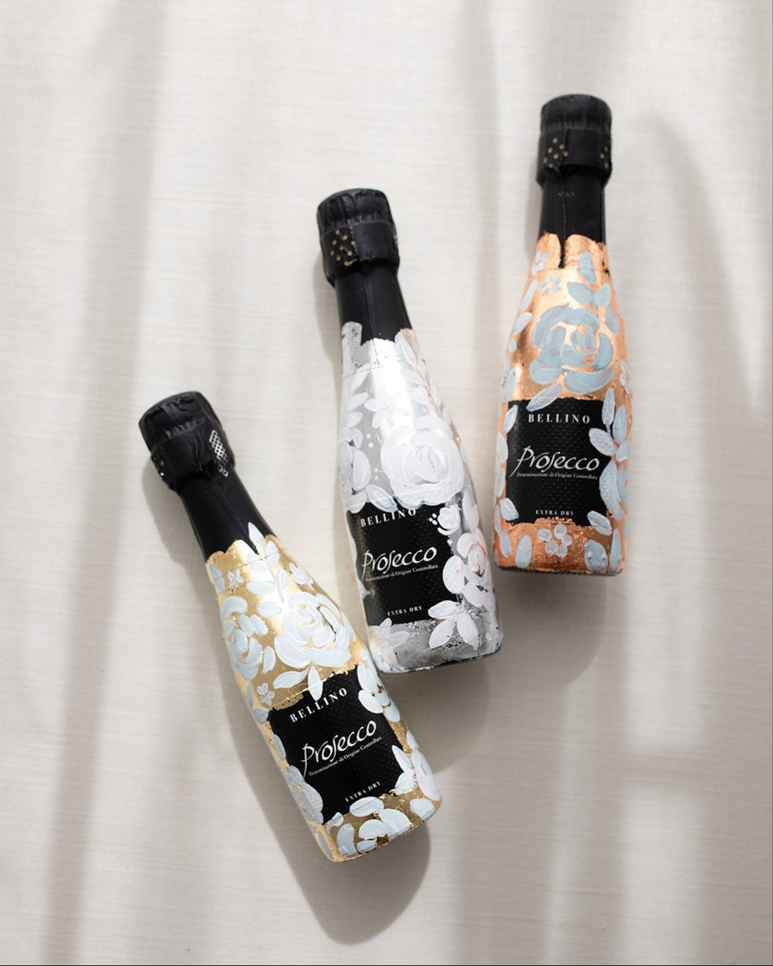 Mini bottles of Prosecco hand painted with a white roses design on a metallic base. Gold, silver and copper examples of the same design. The perfect luxury bridesmaid proposal gift. Fancy Folk Design