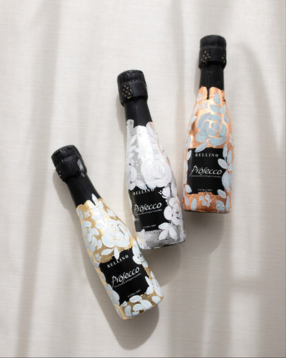 Mini bottles of Prosecco hand painted with a white roses design on a metallic base. Gold, silver and copper examples of the same design. The perfect luxury bridesmaid proposal gift. Fancy Folk Design