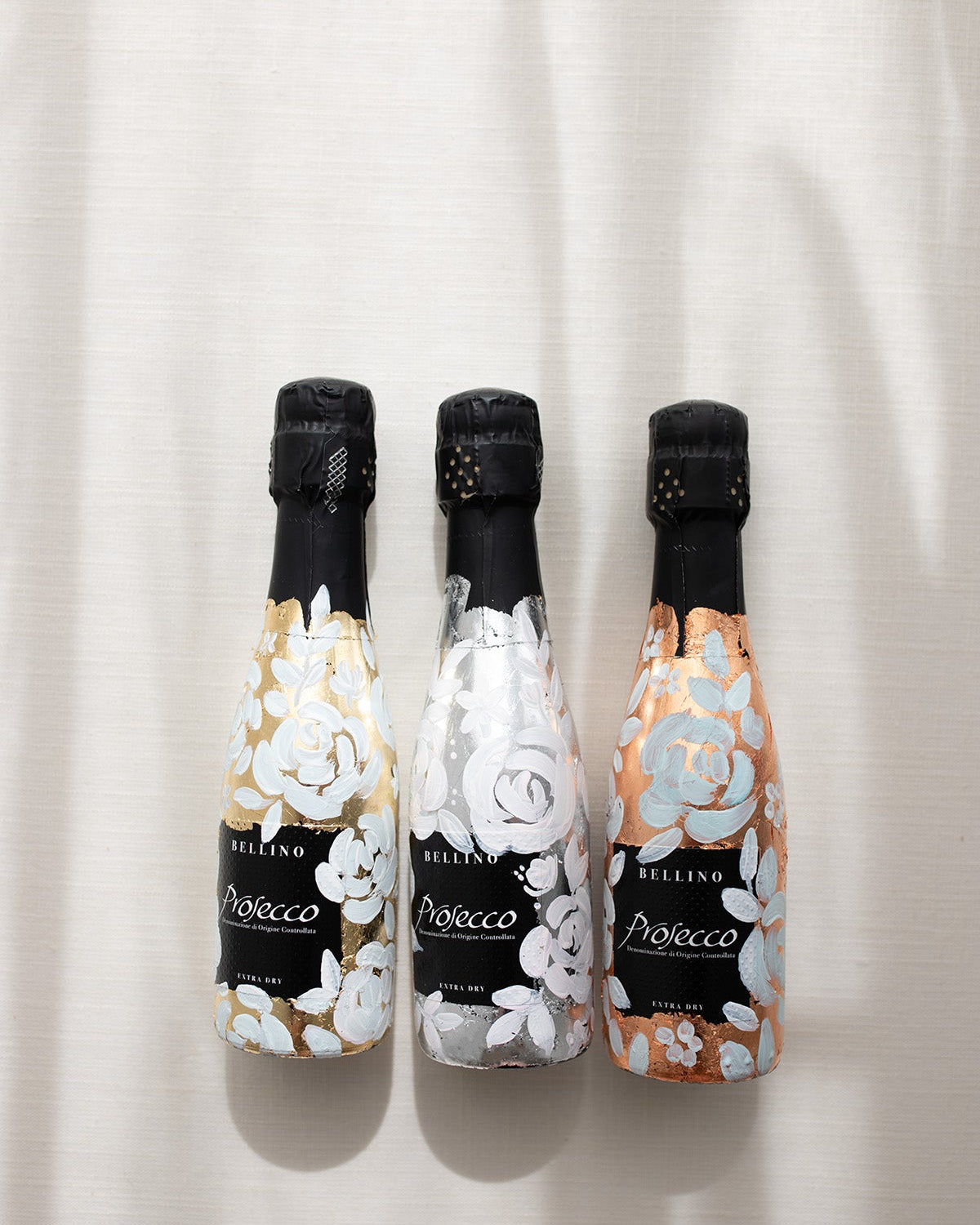 Mini bottles of Prosecco hand painted with a white roses design on a metallic base. Gold, silver and copper examples of the same design. The perfect luxury bridesmaid proposal gift. Fancy Folk Design