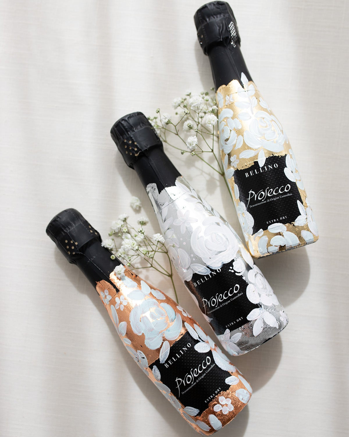 Mini bottles of Prosecco hand painted with a white roses design on a metallic base. Gold, silver and copper examples of the same design. The perfect luxury bridesmaid proposal gift. Fancy Folk Design
