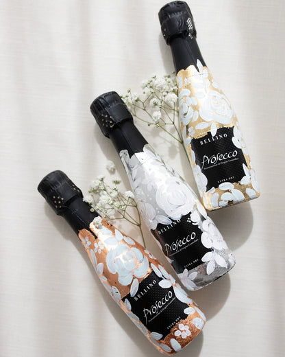 Mini bottles of Prosecco hand painted with a white roses design on a metallic base. Gold, silver and copper examples of the same design. The perfect luxury bridesmaid proposal gift. Fancy Folk Design